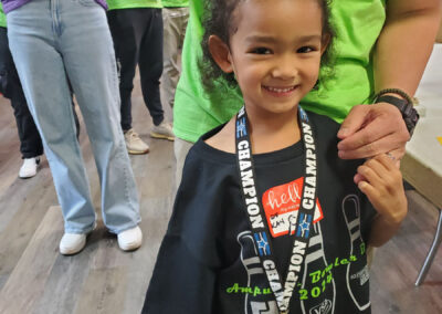 A young girl supporting the Bowler Bash with a t-shirt and a medal, smiling with her parent.